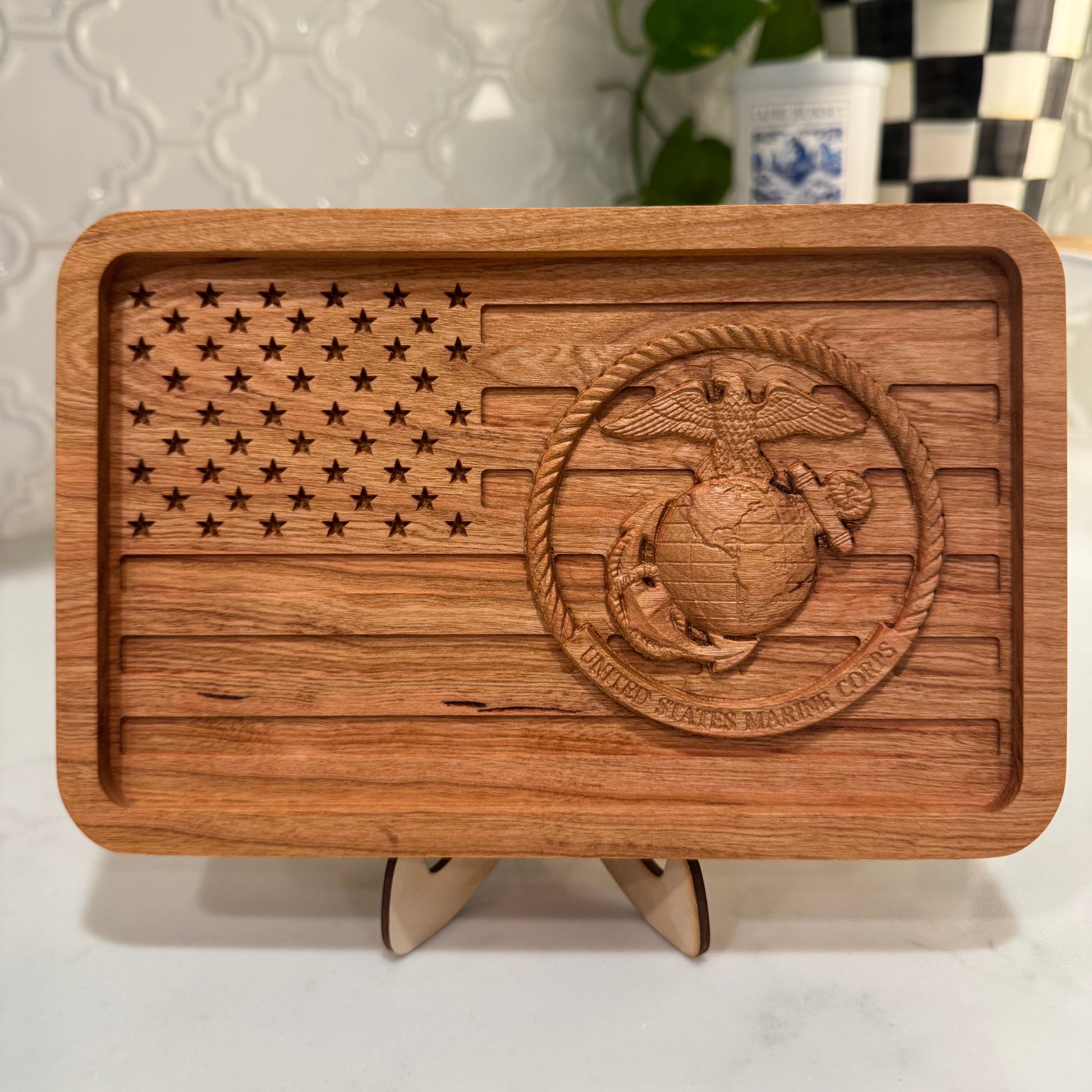 A rectangular wooden tray with rounded corners, featuring a laser-engraved American flag design on the left and a raised Marine Corps emblem on the right, displayed on a small stand against a kitchen countertop background.