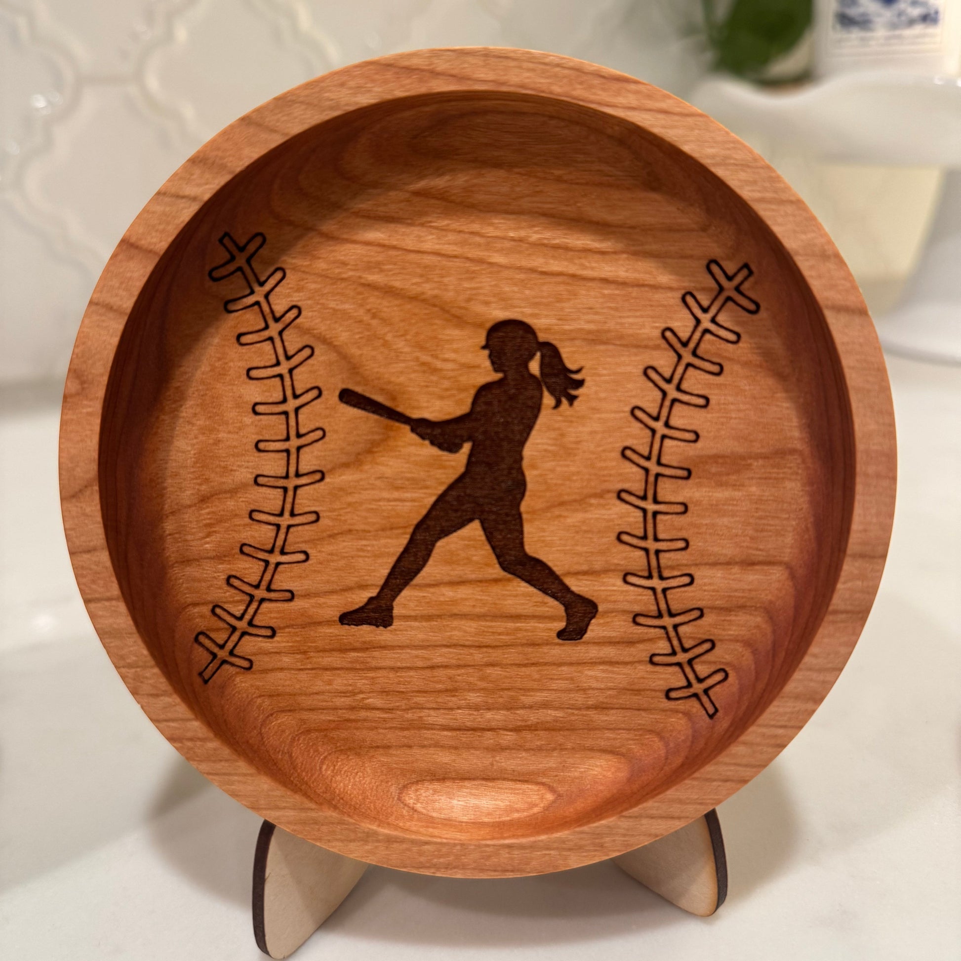 A round wooden bowl with a baseball design, featuring a dark brown engraved silhouette of a softball player in the center and baseball seam lines on both sides. The bowl is displayed on a small wooden stand against a light-colored countertop background.